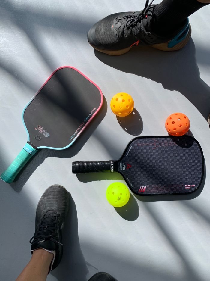 Two pickleball paddles and balls on a sunlit court, with players' feet showing.