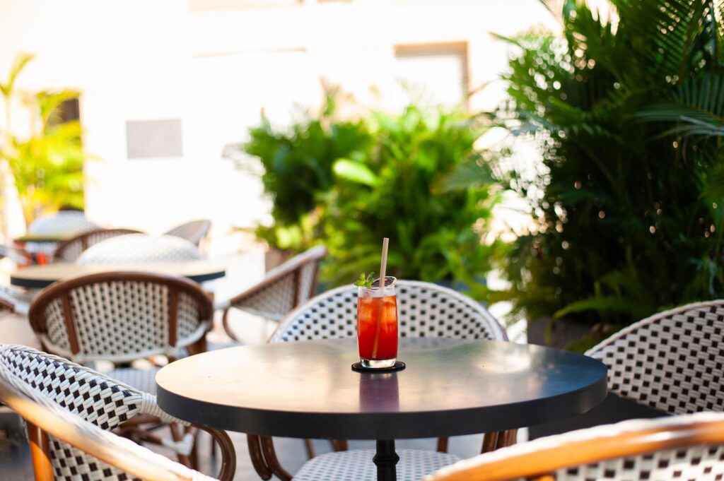 A vibrant cocktail sits on a sunlit patio table surrounded by lush plants, creating a serene outdoor cafe vibe.